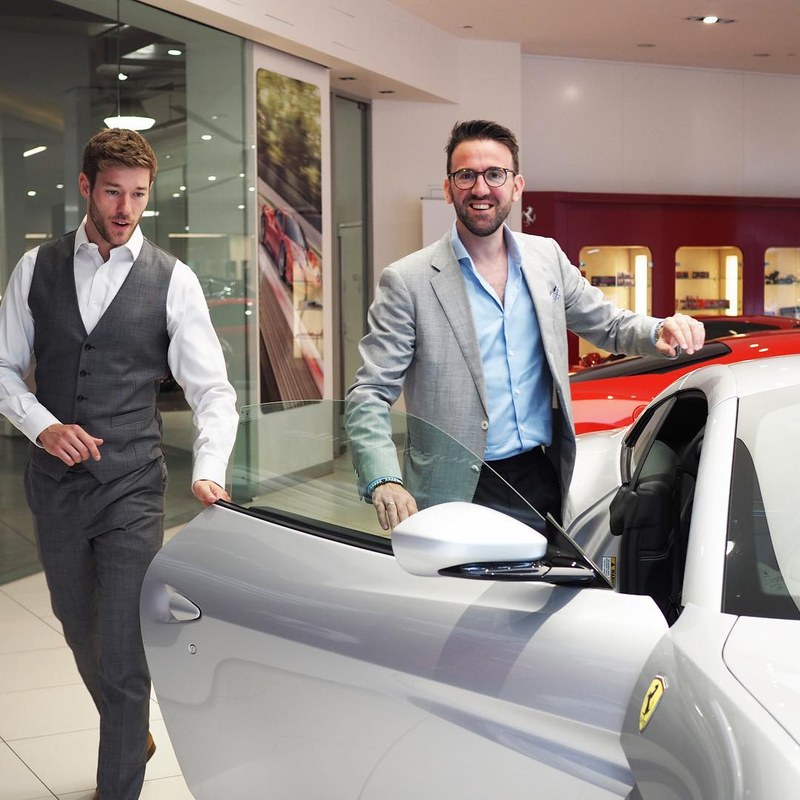 Carl in a grey jacket beside a silver Ferrari in a dealership showroom