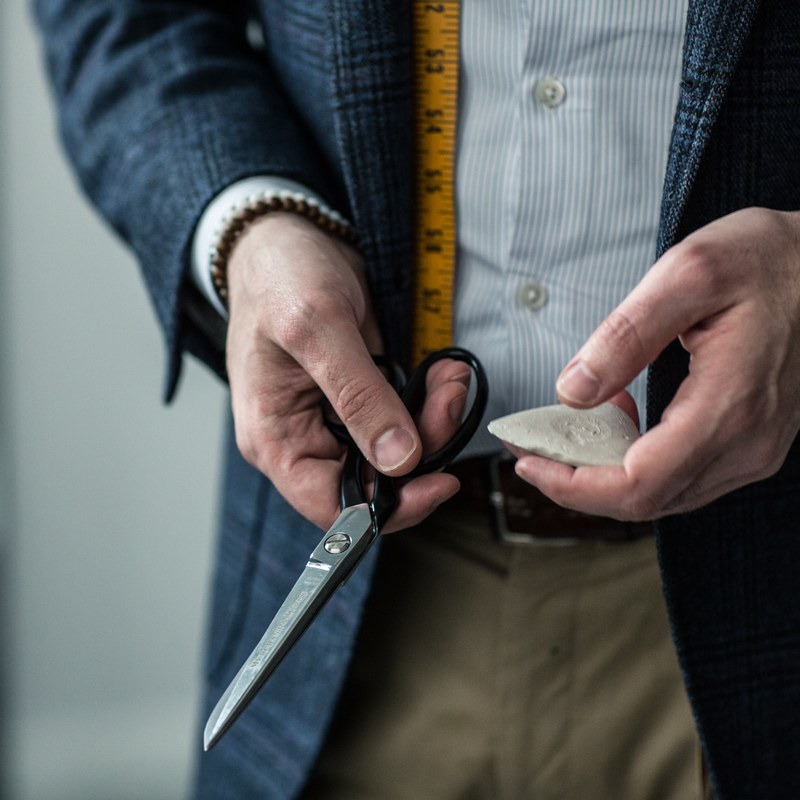 Carl's hands holding tailoring shears and chalk, a tape measure hanging over his shirt