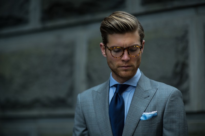 A man in tortoiseshell glasses, a grey check suit and navy tie, looking down