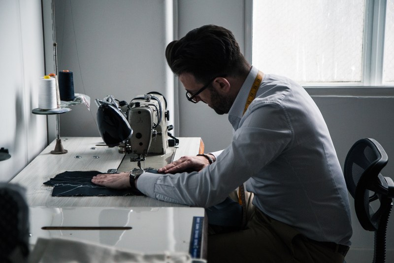Carl at an industrial sewing machine by a window, a tape measure around his neck
